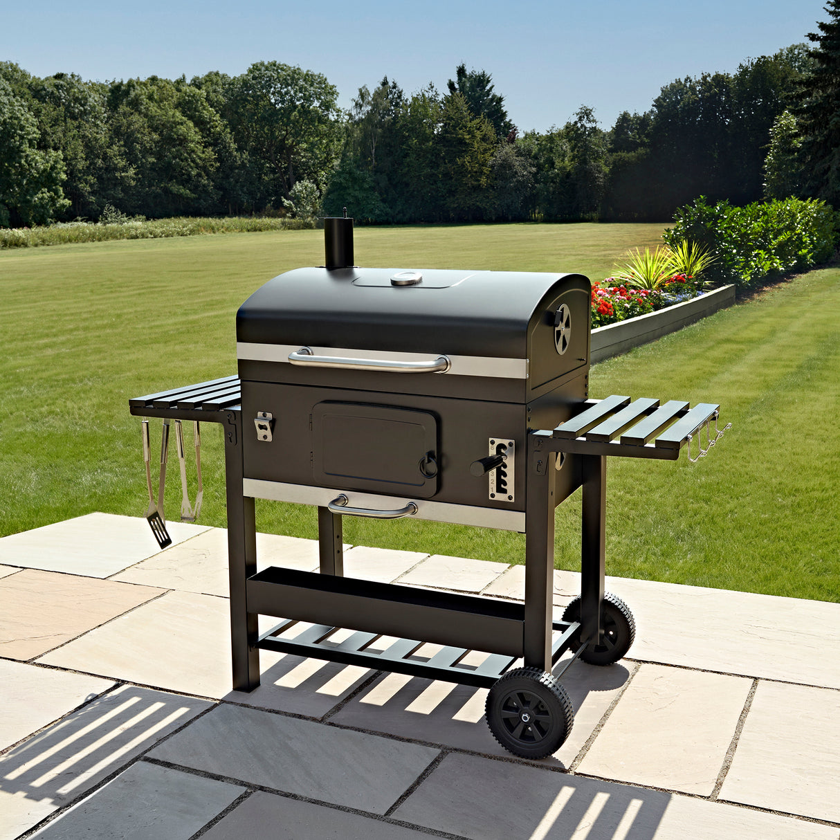 XXL BBQ smoker grill in sleek black with side shelves, wheels for mobility, and a spacious cooking area set on a patio with greenery in the background