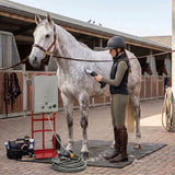 A rider in a helmet grooms a gray horse using a clipper, with grooming tools and a hose nearby in a stable setting