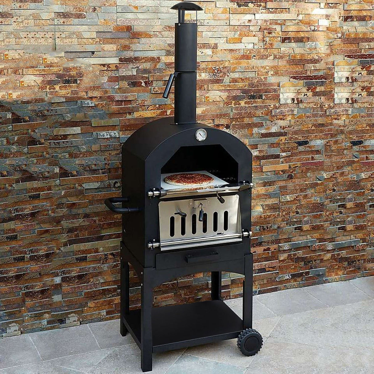 Black KuKoo outdoor pizza oven with stainless steel door, mounted on a sturdy cart, surrounded by a textured stone wall