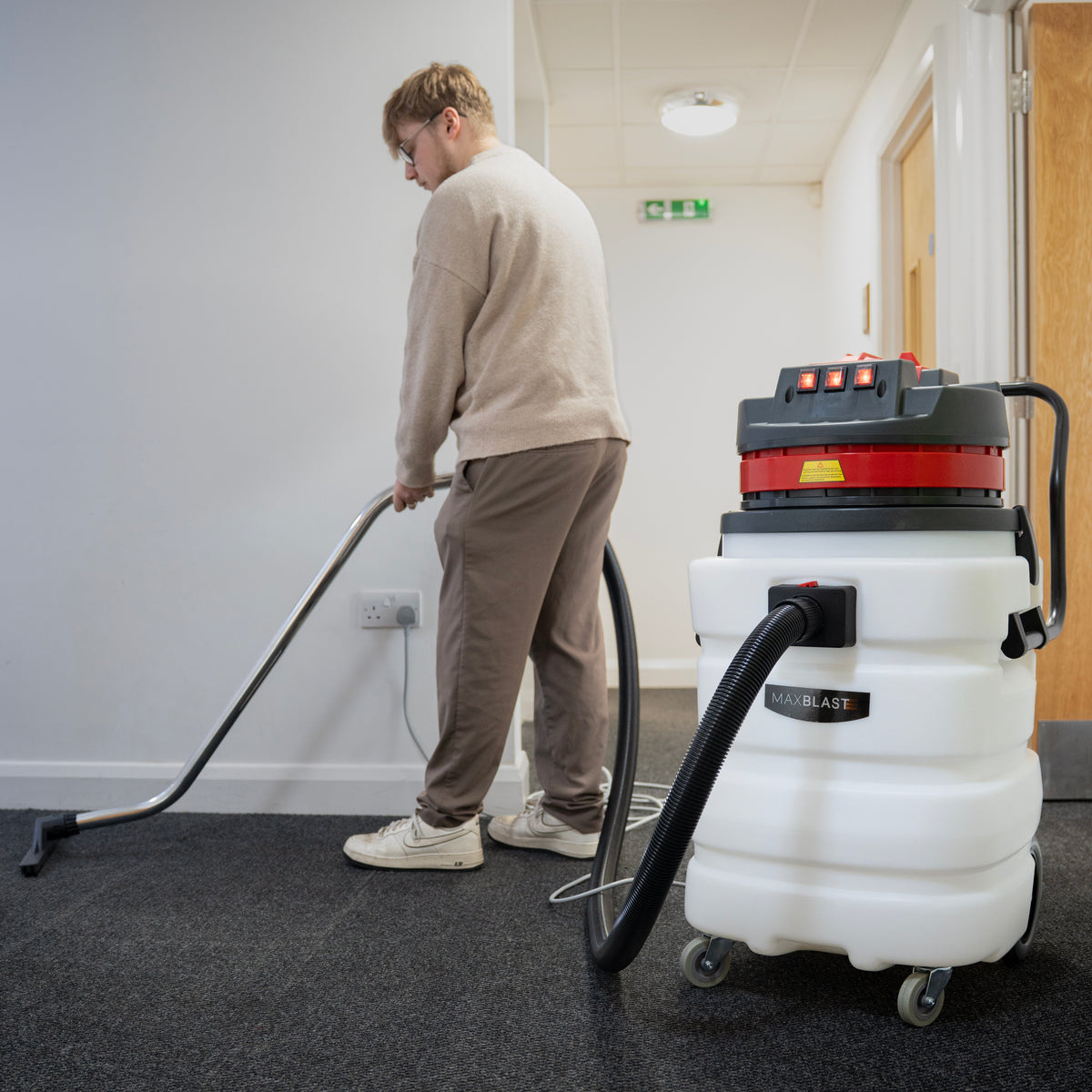 A person using a white and red vacuum cleaner on dark carpet, with a flexible hose and metal wand, in a well-lit hallway