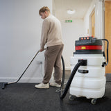 A person using a white and red vacuum cleaner on dark carpet, with a flexible hose and metal wand, in a well-lit hallway