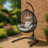 Black hanging rattan egg chair with a cushioned seat, suspended from a sturdy stand, set in a vibrant garden with blooming flowers