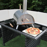 Stainless steel outdoor pizza oven on a table, with a freshly baked pizza on a white plate, surrounded by green grass and a cozy seating area