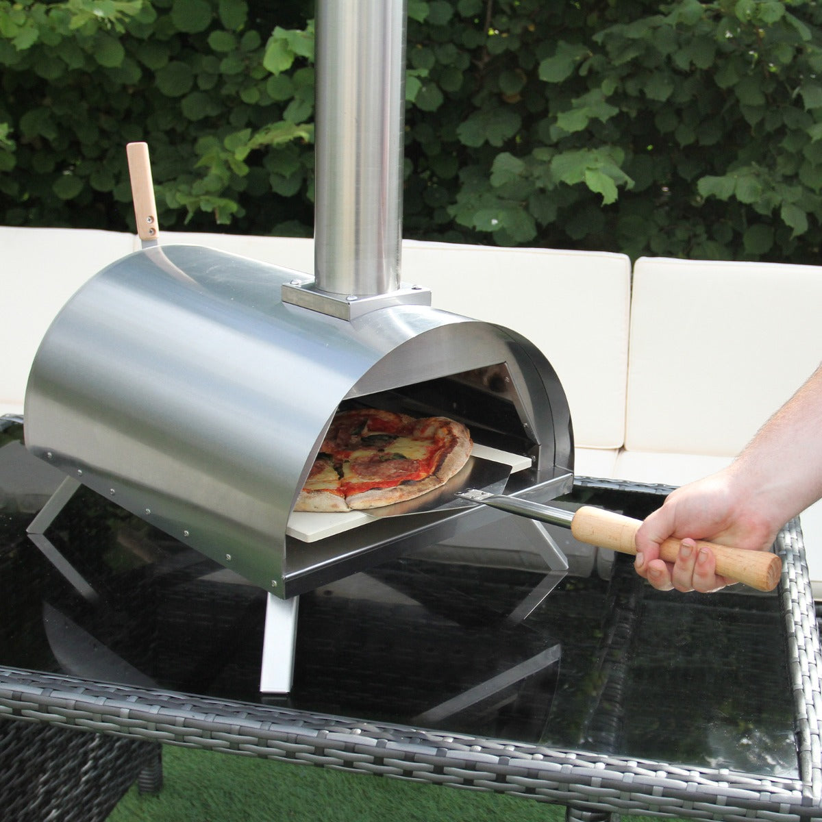 Stainless steel outdoor pizza oven with a sleek dome shape, wooden handles, and a pizza on a stone baking tray, set on a glass table