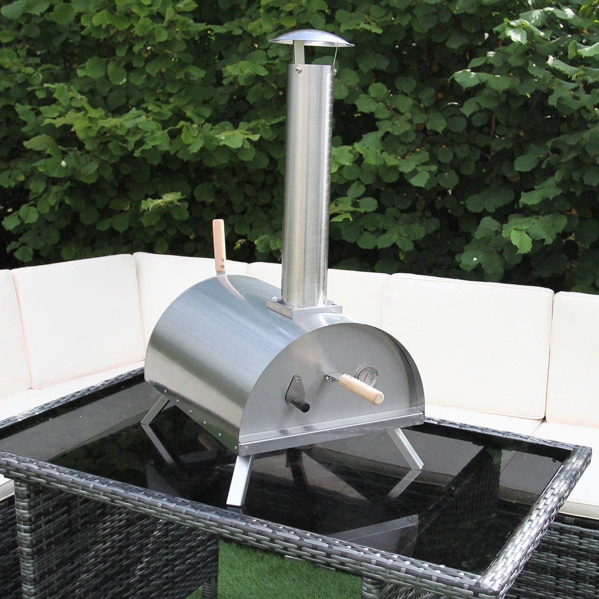 Stainless steel outdoor pizza oven with a tall chimney, set on a glass table surrounded by a cozy seating area and greenery