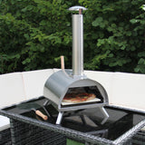 Stainless steel outdoor pizza oven with a chimney, cooking a pizza on a glass-top table surrounded by greenery