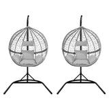 Two grey hanging rattan egg chairs with plush cushions, featuring a sleek black metal stand and a woven, airy design