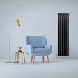 Sleek matt black flat panel radiator stands tall beside a light blue armchair and modern yellow lamp in a minimalist room setting