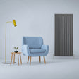 Anthracite grey designer flat panel radiator stands tall beside a light blue armchair and modern yellow floor lamp in a stylish interior setting