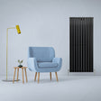 Sleek matt black flat panel radiator stands tall beside a light blue armchair and modern yellow lamp in a minimalist room setting