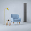 Anthracite grey flat panel radiator stands tall beside a light blue armchair, yellow lamp, and wooden side table with a plant