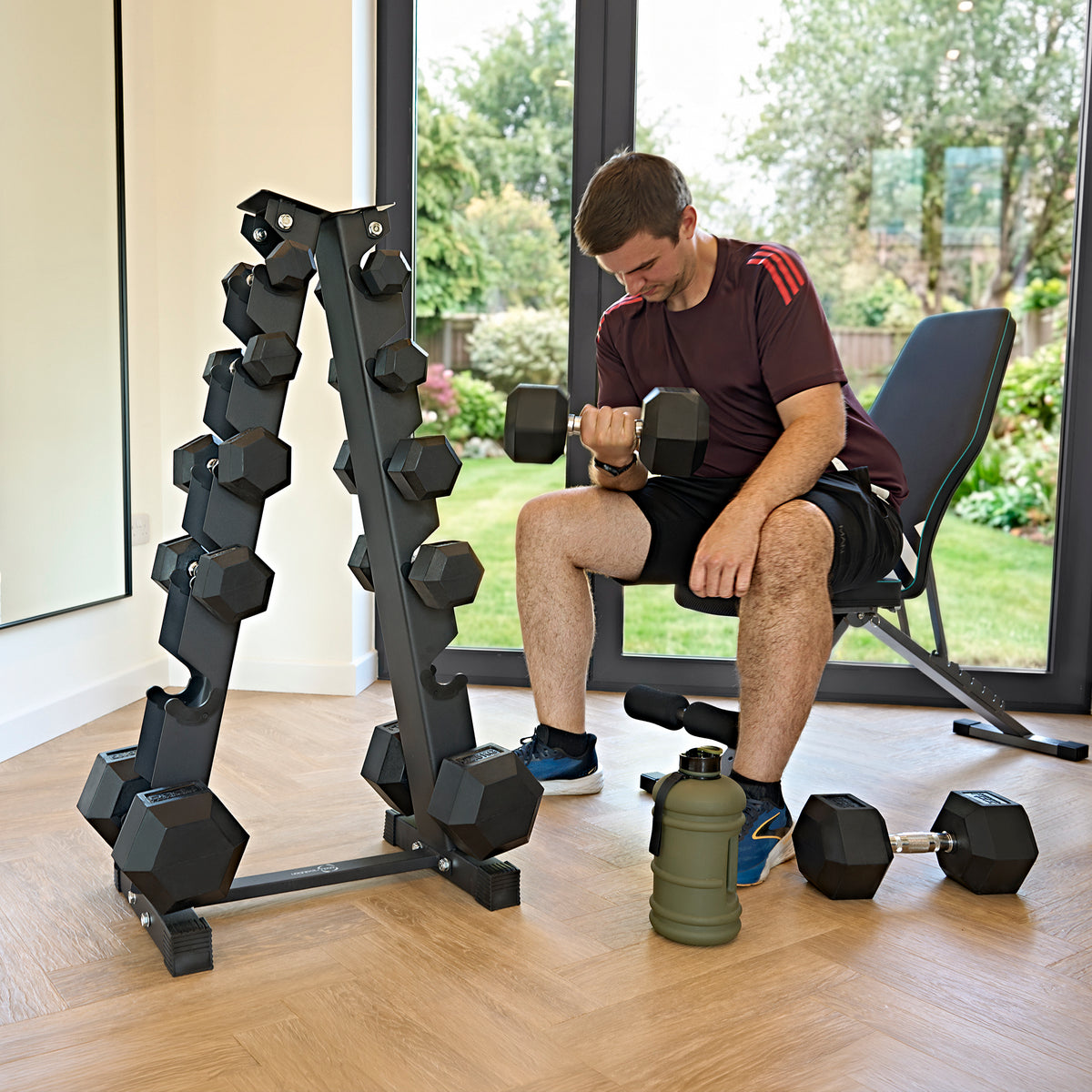 A man in a maroon shirt lifts a dumbbell beside a rack of hexagonal weights in a bright home gym with large windows
