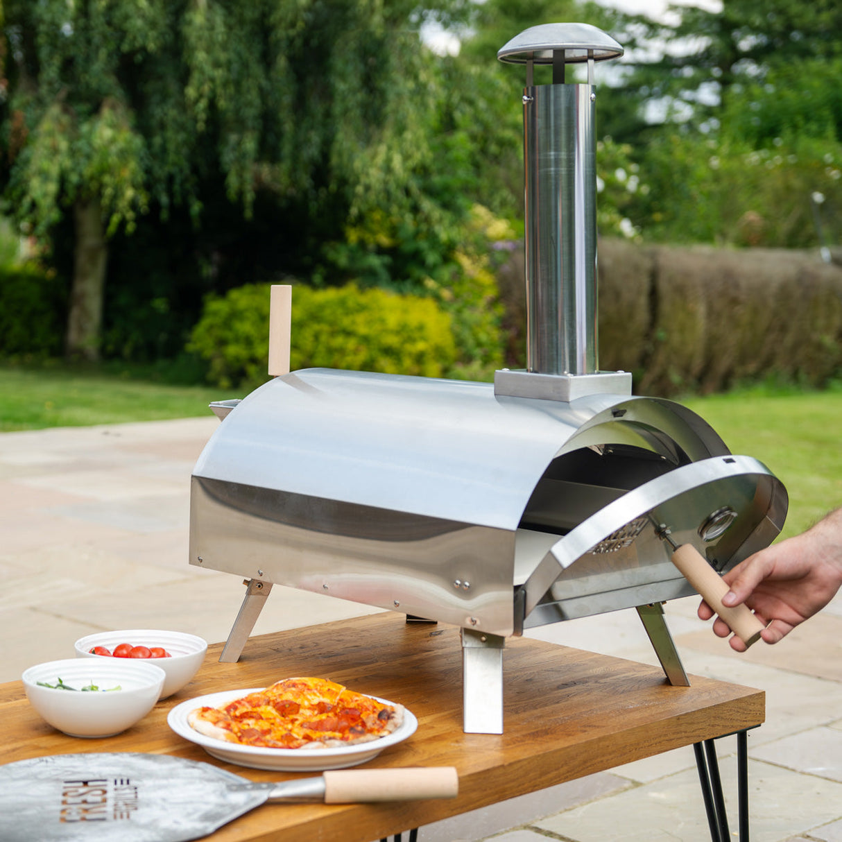 Premium stainless steel outdoor pizza oven with a chimney, set on a wooden table beside fresh pizzas and toppings in bowls