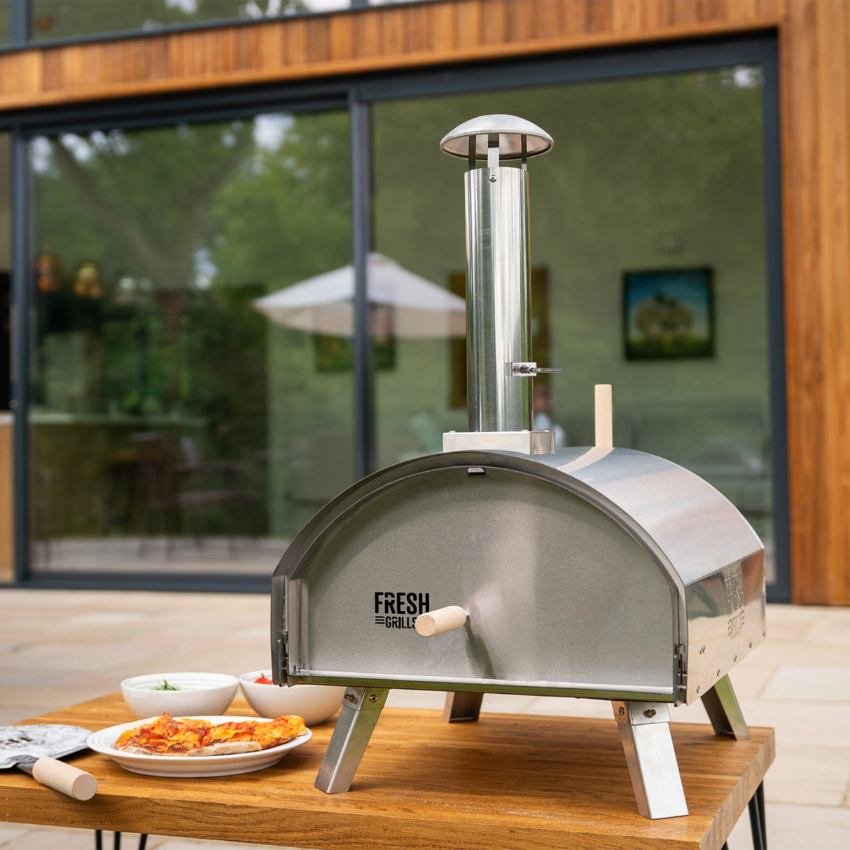 Extra large stainless steel outdoor pizza oven with a sleek dome shape, wooden handles, and a chimney, set on a wooden table