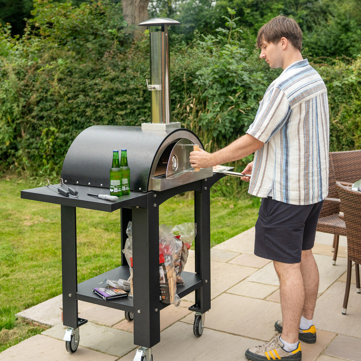 Extra large black pizza oven on a wheeled cart, with a man preparing pizza, surrounded by greenery and outdoor furniture