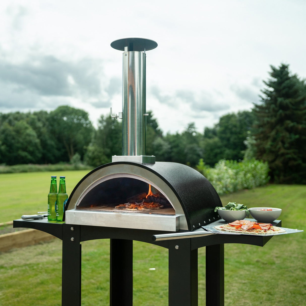 Extra large black pizza oven with stainless steel chimney, surrounded by prep stations, set on a grassy outdoor area with drinks and toppings