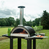 Extra large black pizza oven with stainless steel chimney, surrounded by prep stations, set on a grassy outdoor area with drinks and toppings
