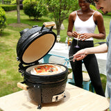 Black 16” Kamado BBQ oven with a textured finish, open lid revealing a pizza inside, set in a sunny outdoor garden scene
