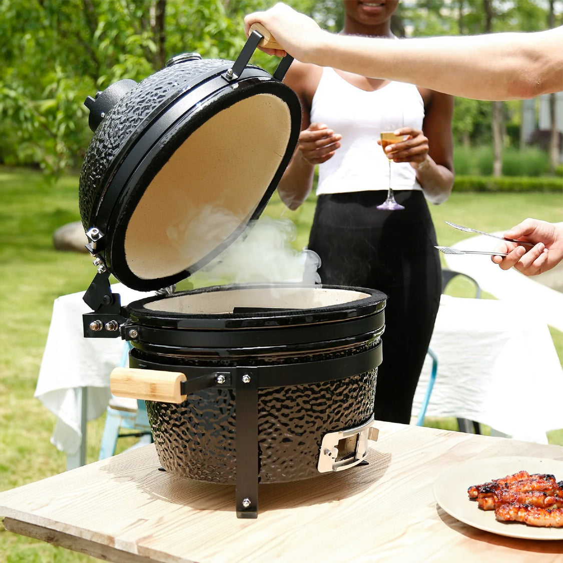 Black 16” Kamado BBQ oven with a textured surface, open lid releasing smoke, surrounded by lush greenery and a person holding a drink