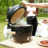 Black 16” Kamado BBQ oven with a textured surface, open lid releasing smoke, surrounded by lush greenery and a person holding a drink