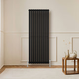 Sleek black oval single column radiator with vertical slats, positioned against a light wall, complemented by stylish valves and a wooden table nearby