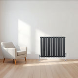 Anthracite grey flat single column radiator with vertical slats, positioned beside a modern white armchair in a bright room