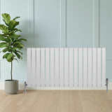Sleek white flat single column radiator with chrome valves, positioned beside a potted plant on a light wooden floor against a pale wall
