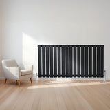Sleek black flat single column radiator with polished valves, positioned beside a modern white armchair in a bright room