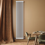 Tall white traditional 4 column radiator with valves, mounted on a warm-toned wall, beside a wooden dining table and soft curtains