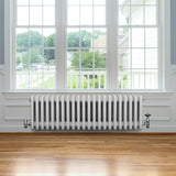 Traditional white 4 column radiator with valves, featuring a sleek design, installed beneath a large window in a bright room with wooden flooring