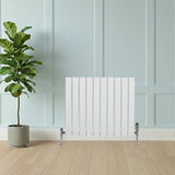 Sleek white flat single column radiator with vertical slats, mounted on a light wood floor against a soft blue wall, complemented by a green plant
