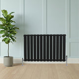 Sleek black flat single column radiator with vertical slats, positioned beside a potted plant on a light wood floor against a pale wall