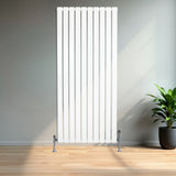 Sleek white flat single column radiator with vertical slats, standing on chrome feet, set against a modern interior with a potted plant