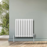 Sleek white oval single column radiator with vertical bars, mounted on a wall in a bright room with wooden flooring and greenery outside