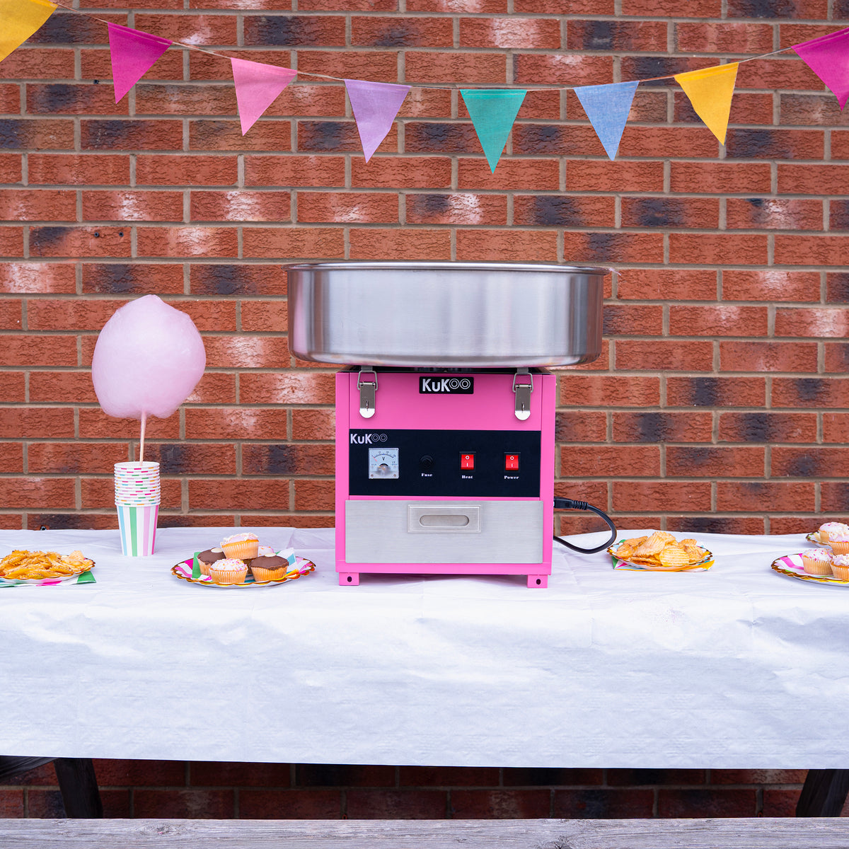 Cotton candy machine in pink with stainless steel bowl, set against a brick wall, surrounded by colorful bunting and party treats