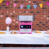 Cotton candy machine in pink with stainless steel bowl, set against a brick wall, surrounded by colorful bunting and party treats