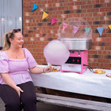 A woman in a pink blouse smiles while holding a large fluffy cotton candy, next to a pink cotton candy machine and colorful bunting
