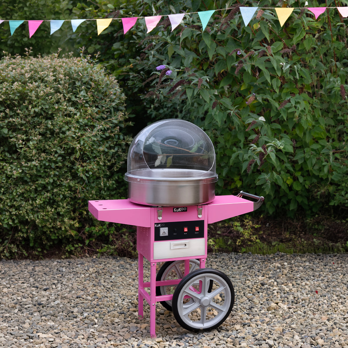 Bright pink cotton candy machine on a wheeled cart, featuring a clear dome and silver bowl, set against a lush green backdrop