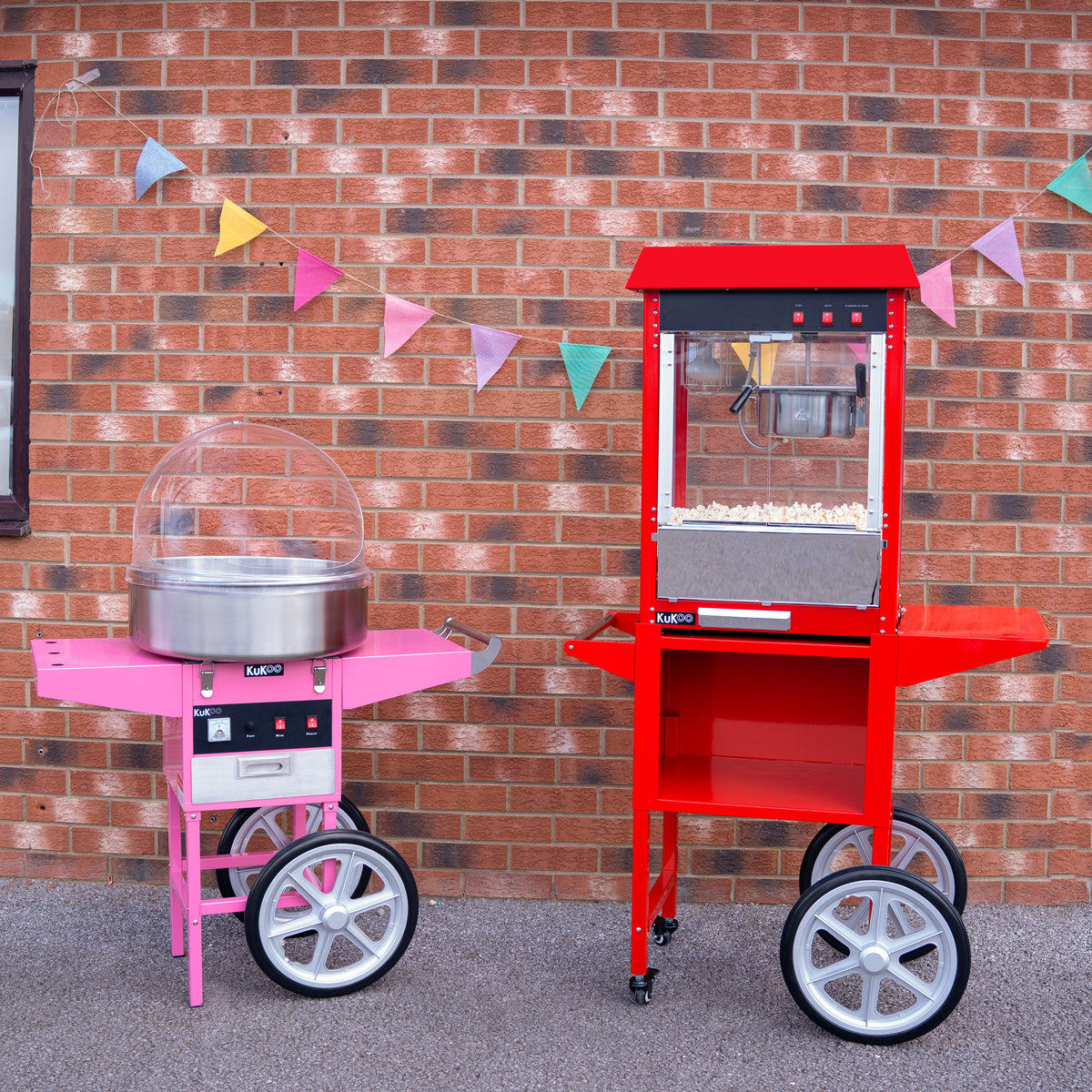 Bright pink cotton candy machine and vibrant red popcorn machine on wheels, set against a brick wall with colorful bunting