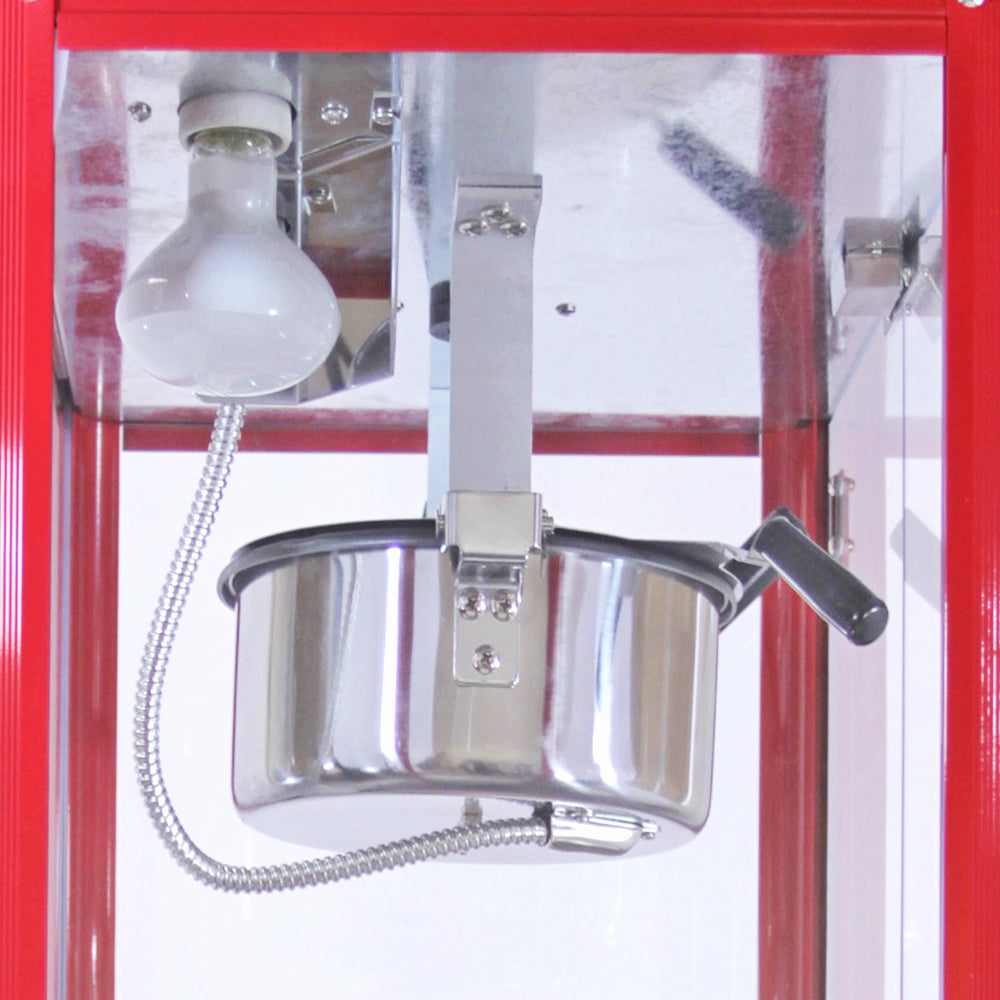 Shiny stainless steel popcorn kettle with a handle, mounted under a bright light in a red popcorn machine cabinet