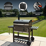 Black outdoor grill with a front removable shelf, rain cover, and wire cleaning brush, set on a patio with green grass in the background