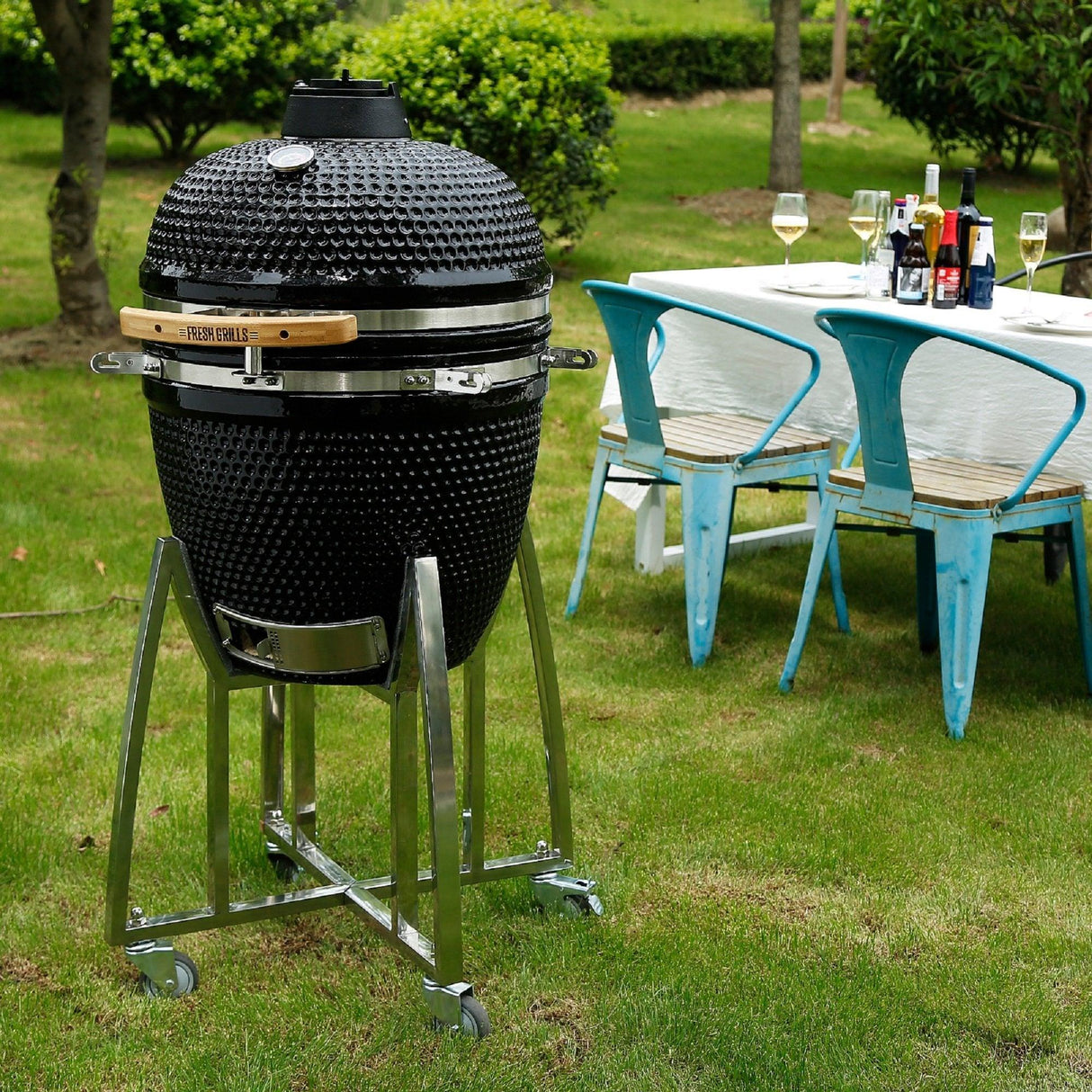Black ceramic Kamado charcoal BBQ grill on a stainless steel cart, set in a green outdoor space with a dining table nearby