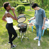 Two people enjoying a barbecue outdoors, with a black Kamado charcoal grill and sizzling steaks on the grill, surrounded by greenery