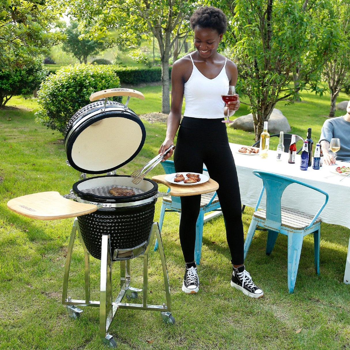Kamado charcoal BBQ grill in black with wooden side shelves, a woman grilling outdoors, surrounded by greenery and a dining setup